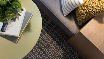 Una habitación con un tapete estampado, una mesa redonda con un libro y una planta, un sofá con un cojín a rayas y una manta con textura.
