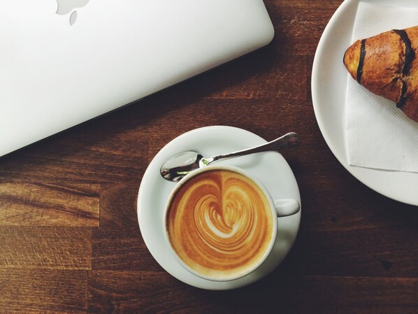 A cup of coffee with latte art on a saucer, a spoon, a laptop, and a plate with a croissant on a wooden table.