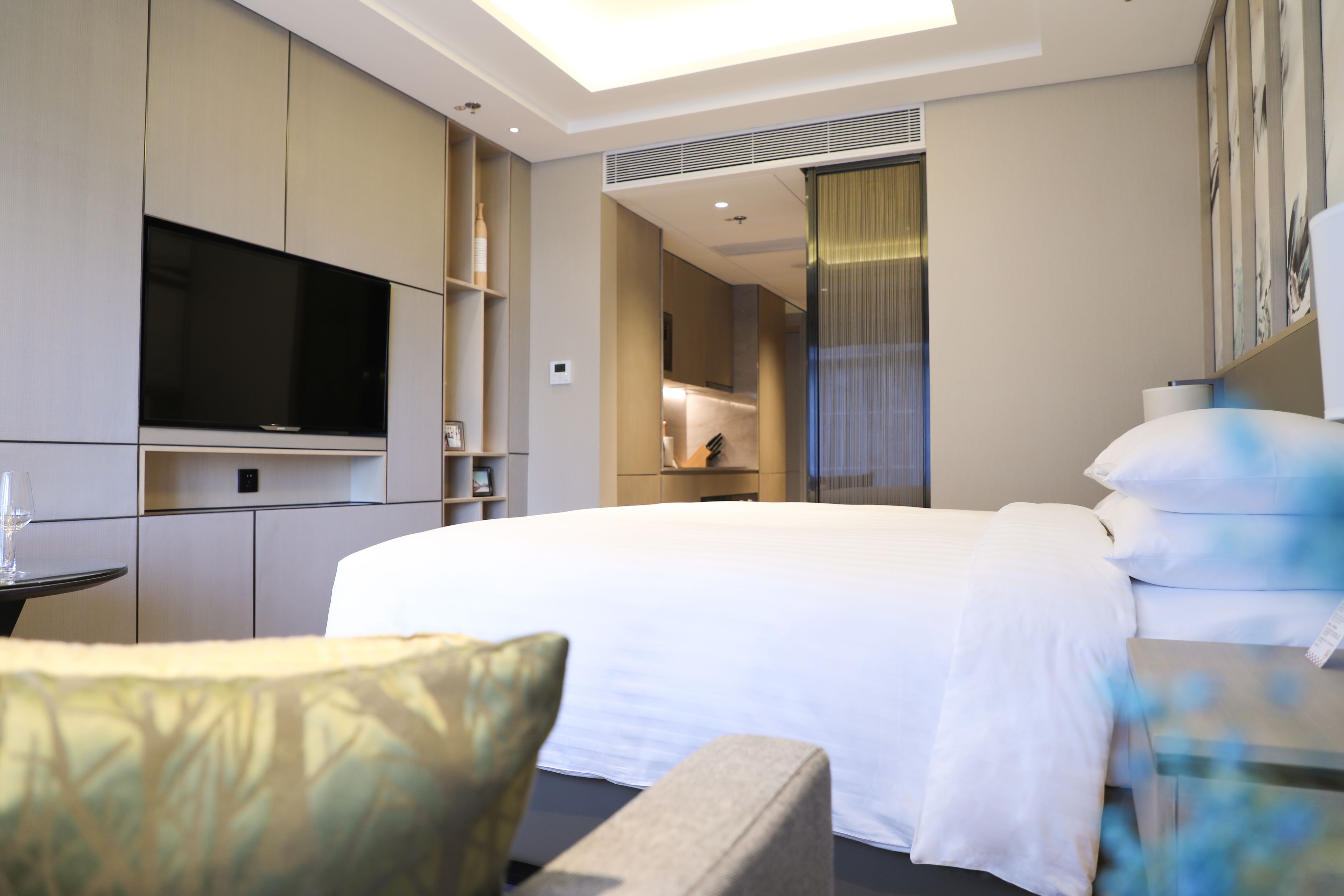 A modern hotel room with a large bed, a flat-screen TV mounted on the wall, a small table with a glass, and a chair.