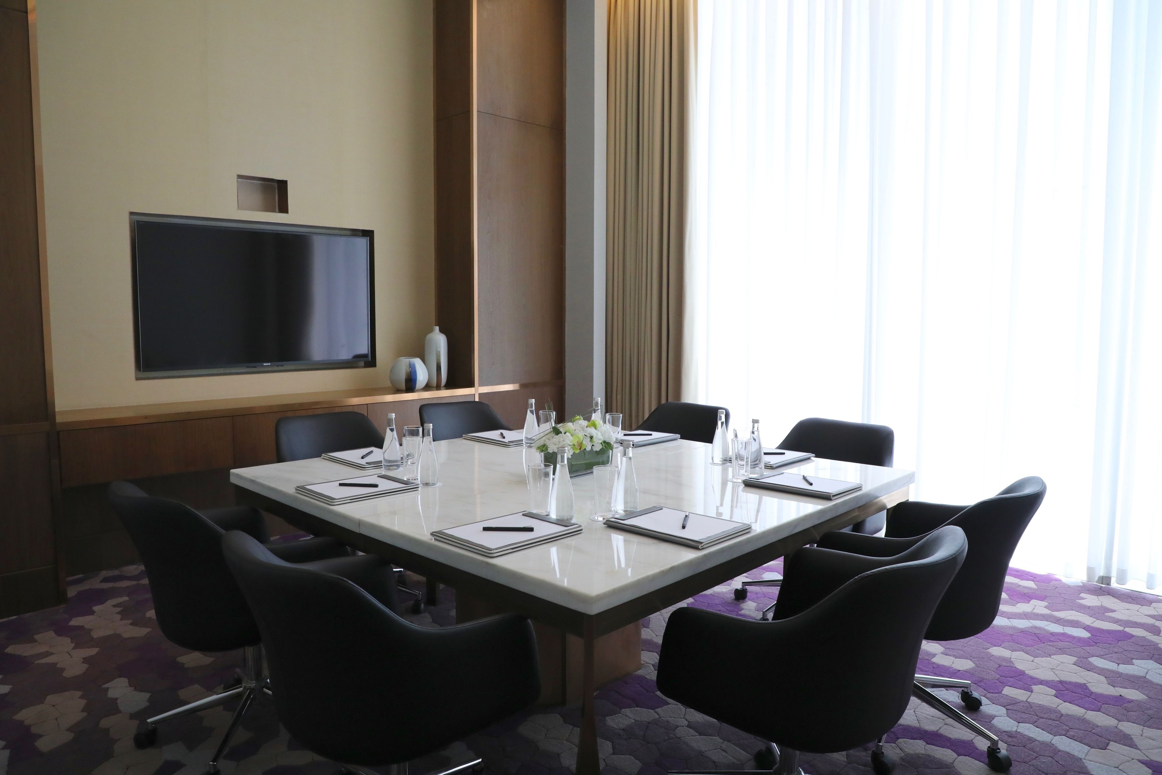 A conference room with a large table, six chairs, a flat-screen TV, and a window with curtains.