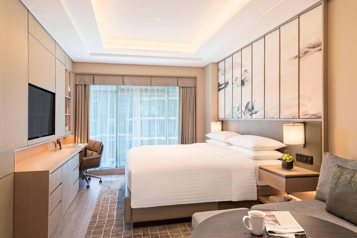Een hotelkamer met een groot bed, een bureau met stoel, een televisie, een nachtkastje met lamp en een raam met gordijnen.