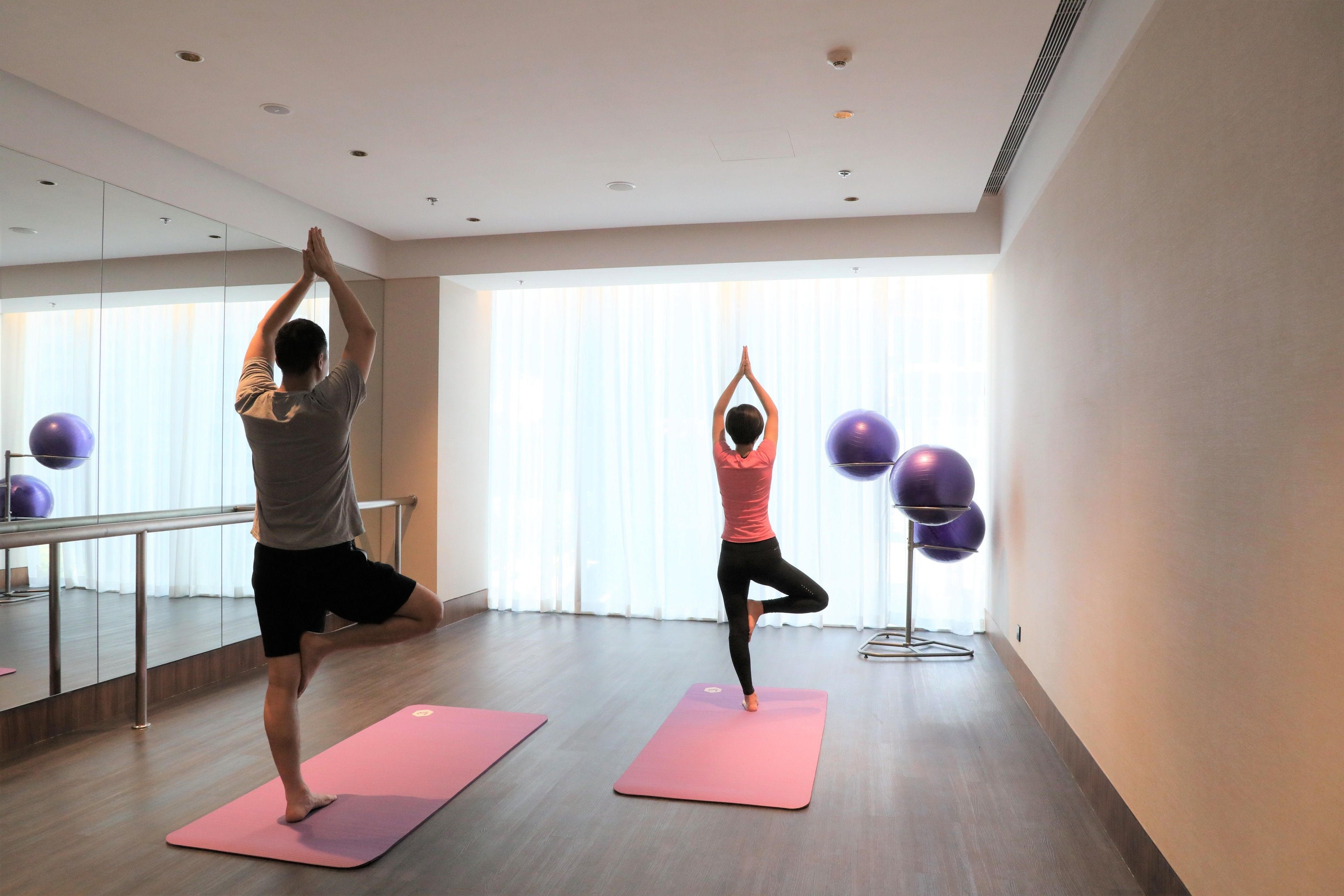 Ein Yogastudio mit zwei Personen, die auf rosafarbenen Matten üben, einer Spiegelwand und Trainingsbällen.