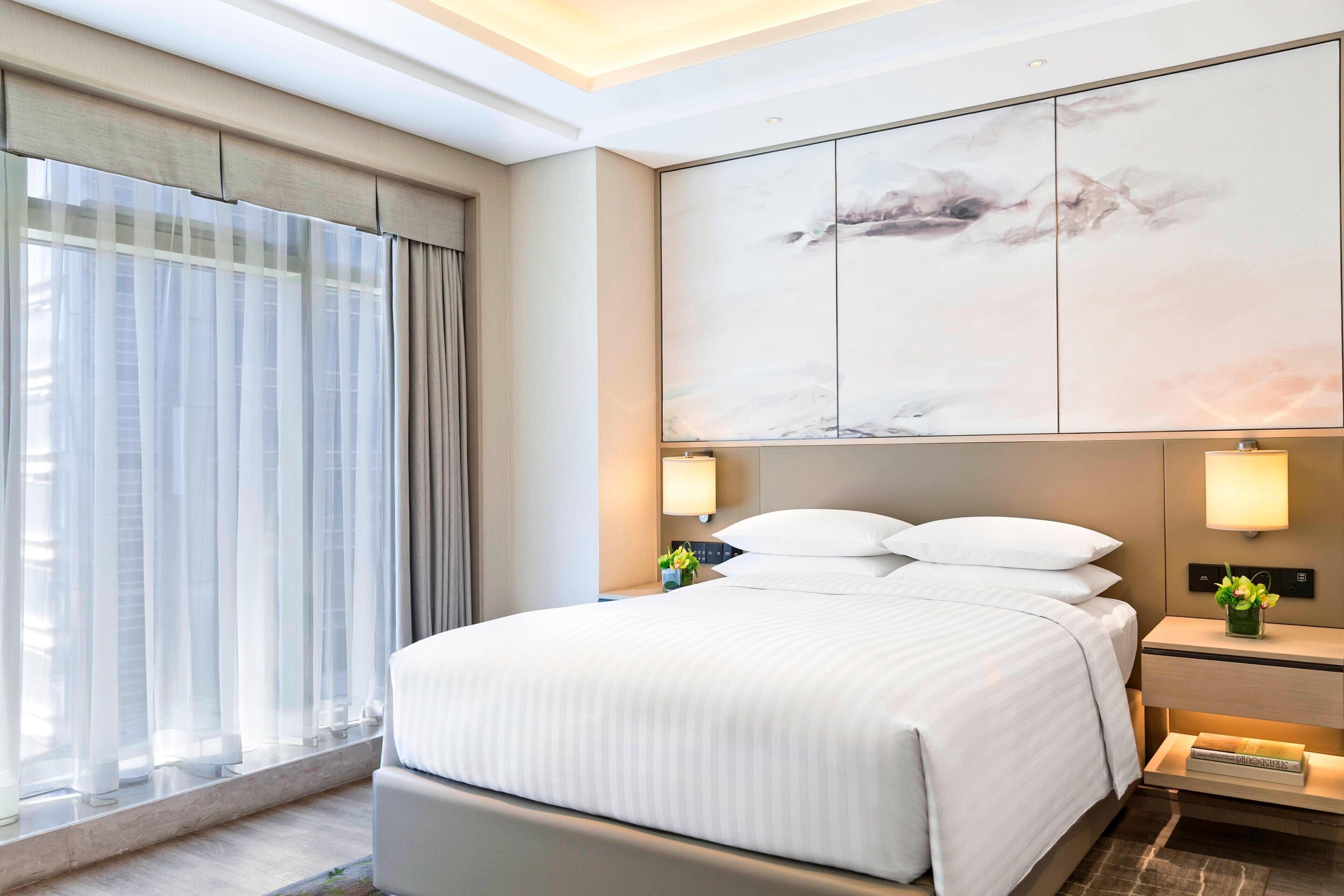 Een hotelkamer met een groot bed, een nachtkastje met een lamp, een aan de muur bevestigde televisie en een raam met dunne gordijnen.