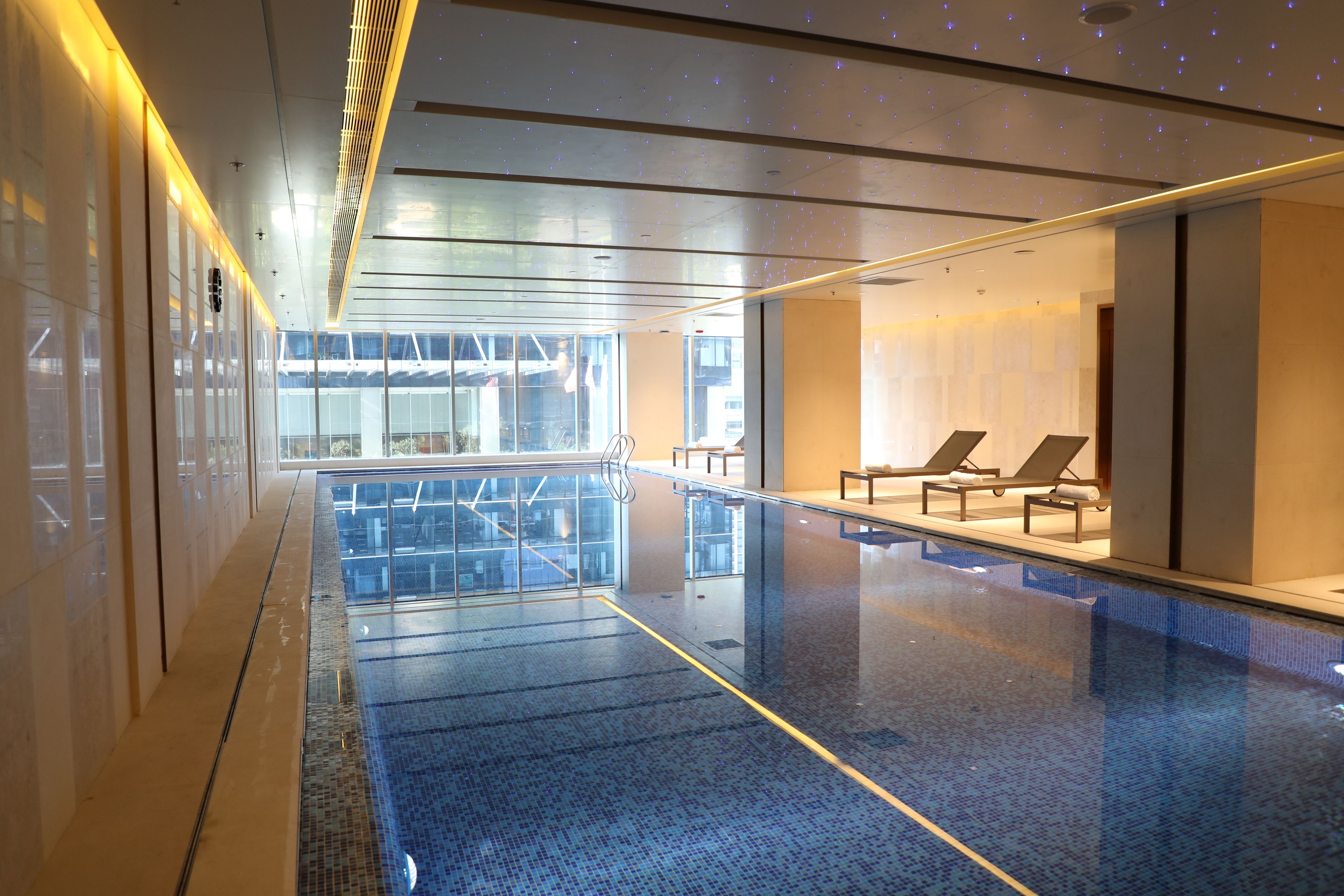 Una piscina interior moderna con paredes de vidrio transparente, sillas de descanso y vistas al exterior.