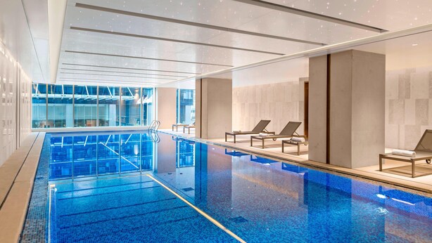 Piscina interior moderna com água azul cristalina, cadeiras de descanso e uma parede de vidro que oferece vista para o exterior.
