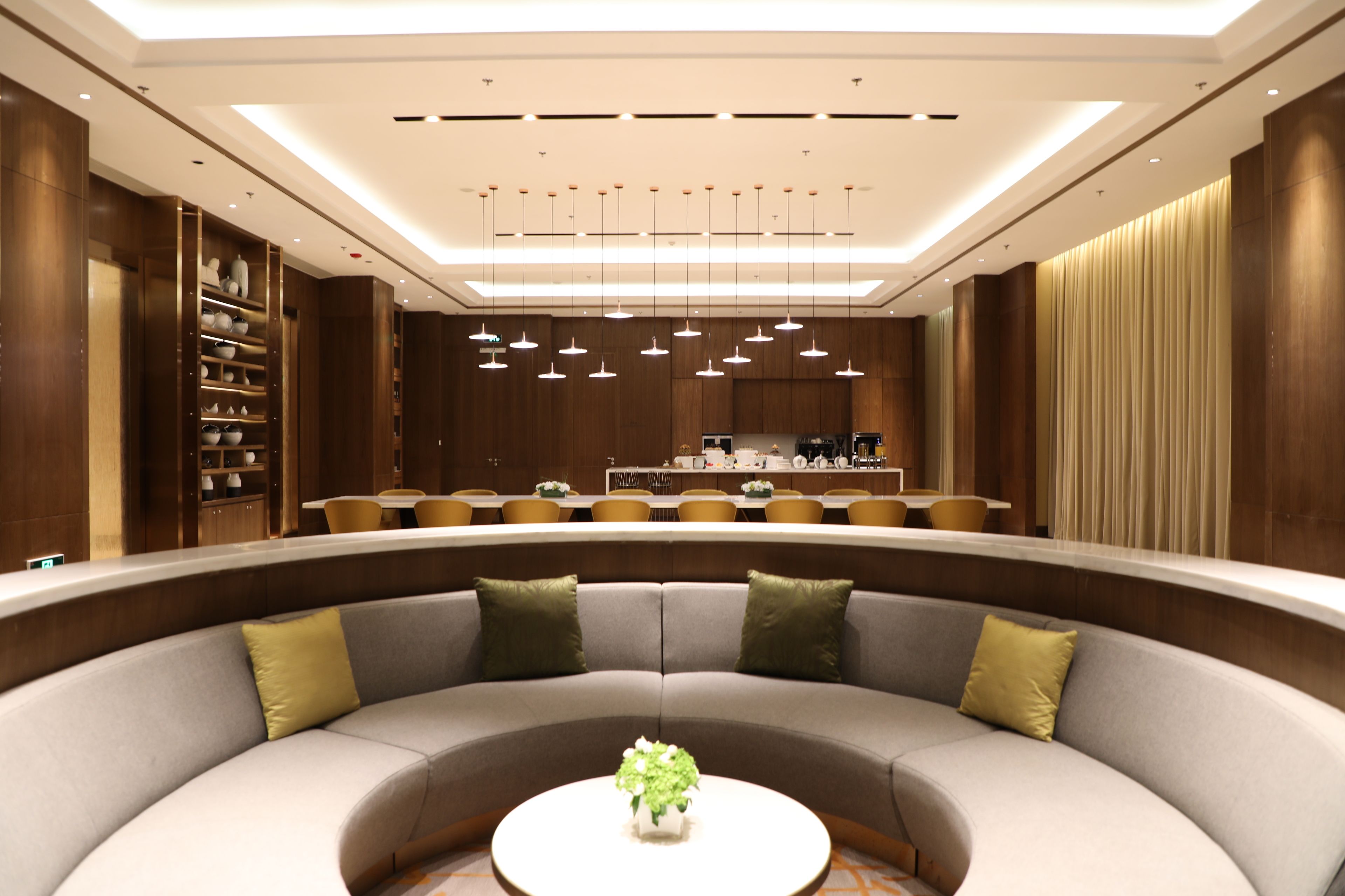 A modern hotel lobby with a curved seating area, a central coffee table, and a kitchen in the background.