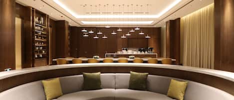 A modern hotel lobby with a curved seating area, a central coffee table, and a kitchen in the background.