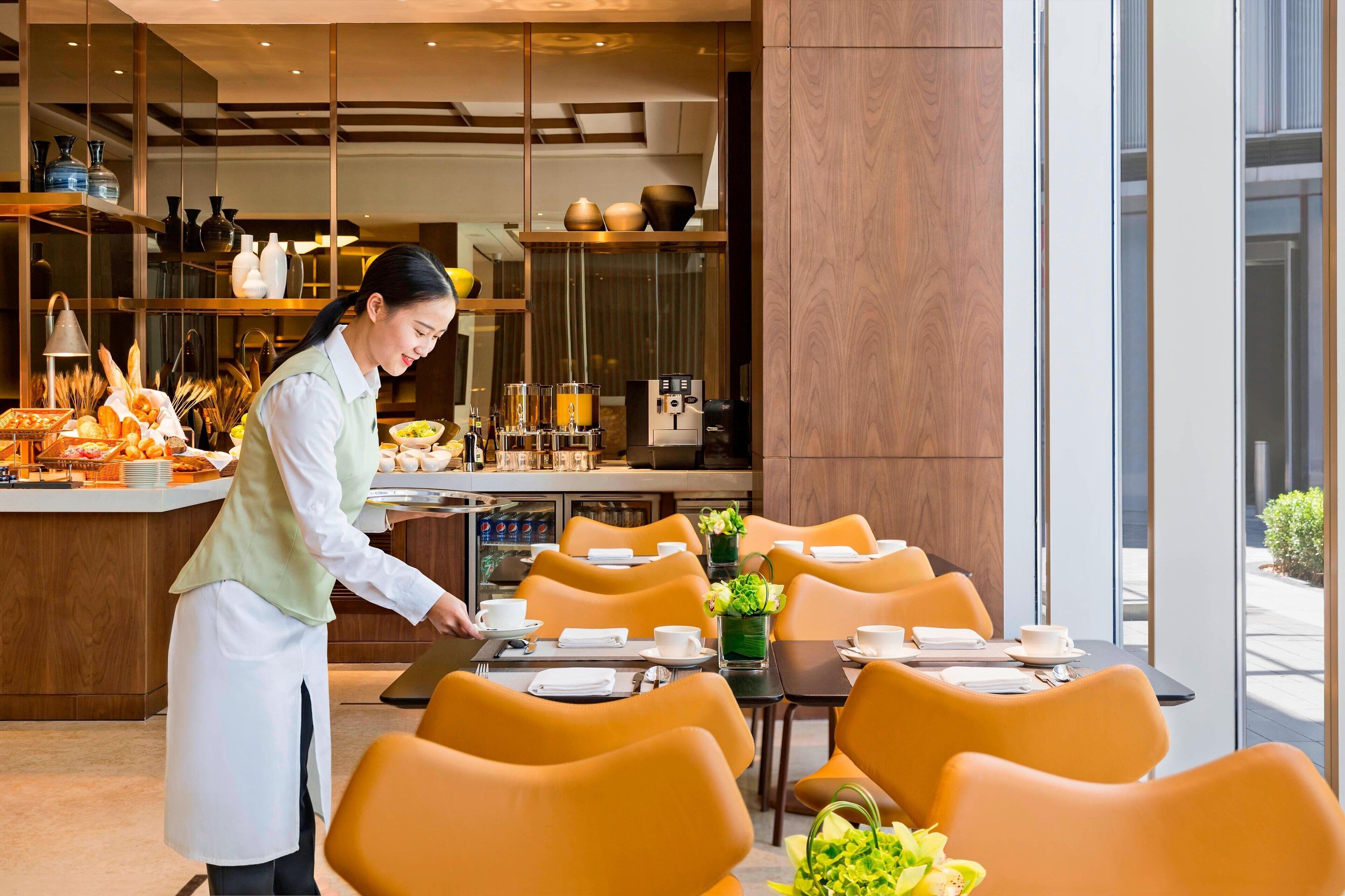 A modern restaurant interior with a buffet setup, orange chairs, and a waiter serving.