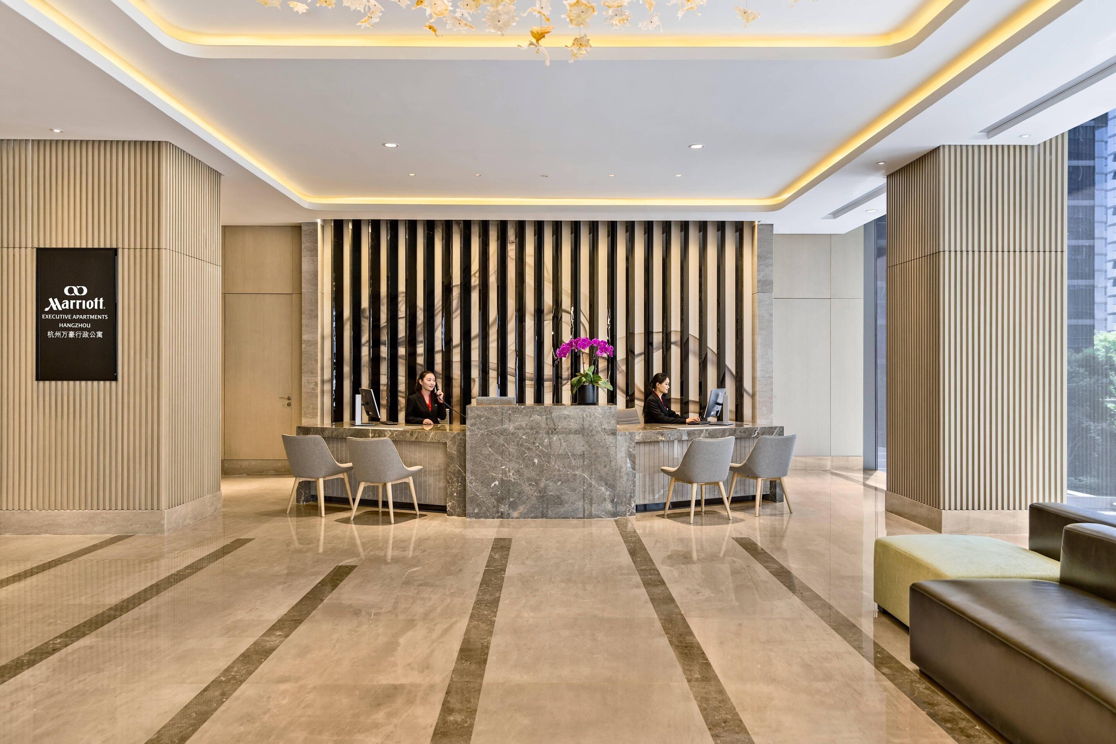A modern hotel reception area with a marble reception desk, two receptionists, and a large window providing a view of the city.