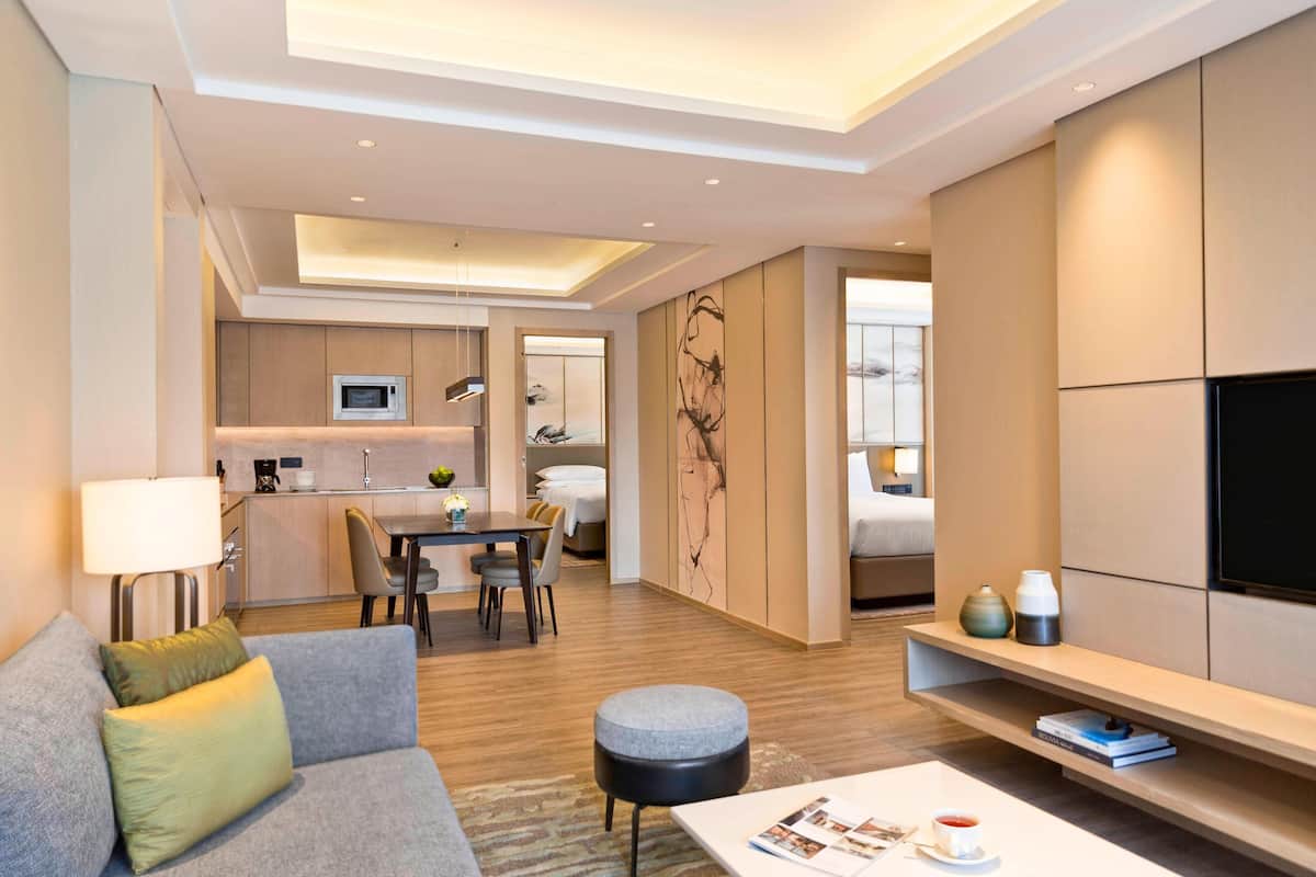 Een moderne hotelkamer met een grijze bank, een salontafel, een flatscreen-televisie, een zithoek met een tafel en stoelen, en een keukenhoek met een magnetron.