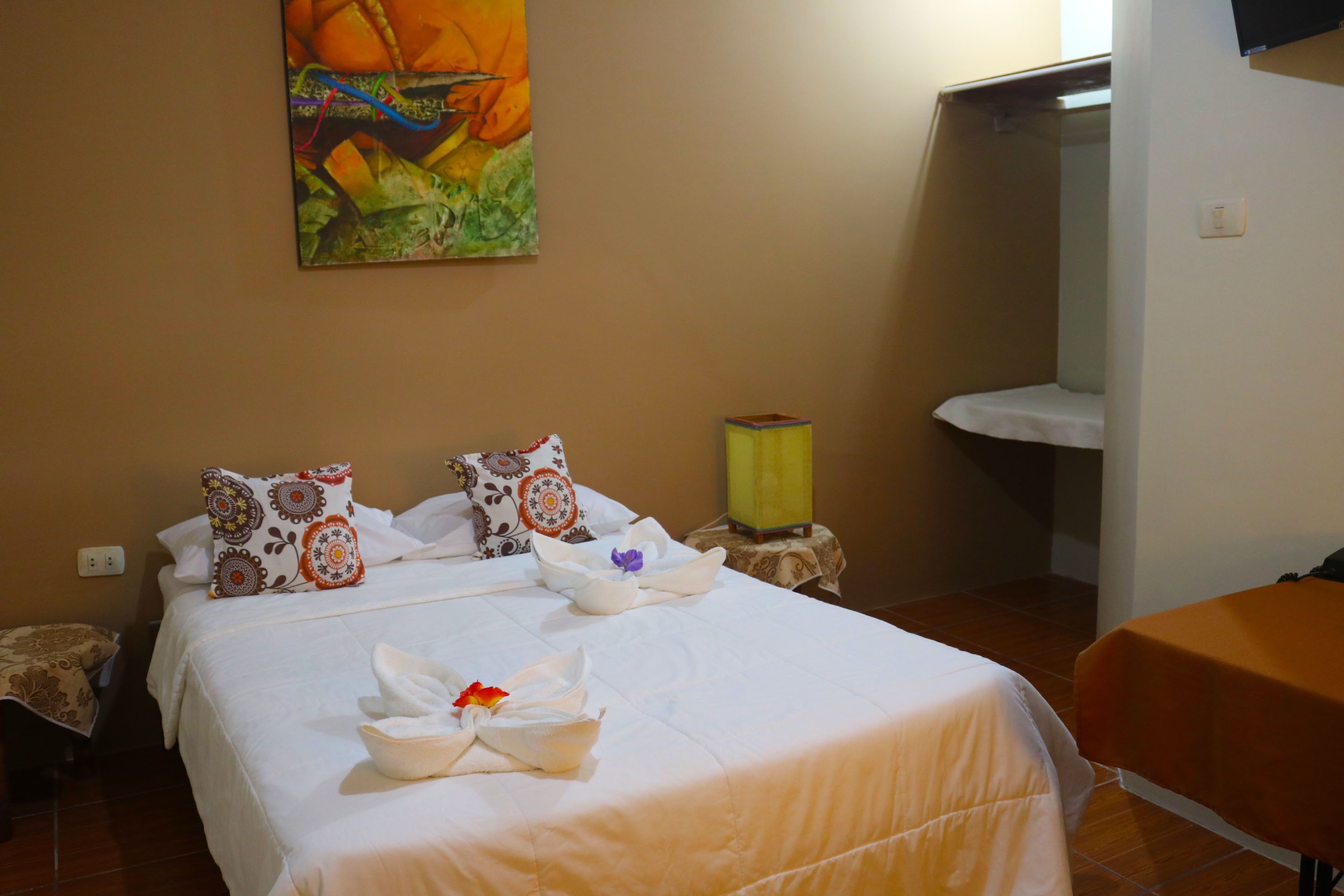 A hotel room with a bed, pillows, a painting, a bedside table with a lamp, and a small table with a tablecloth.