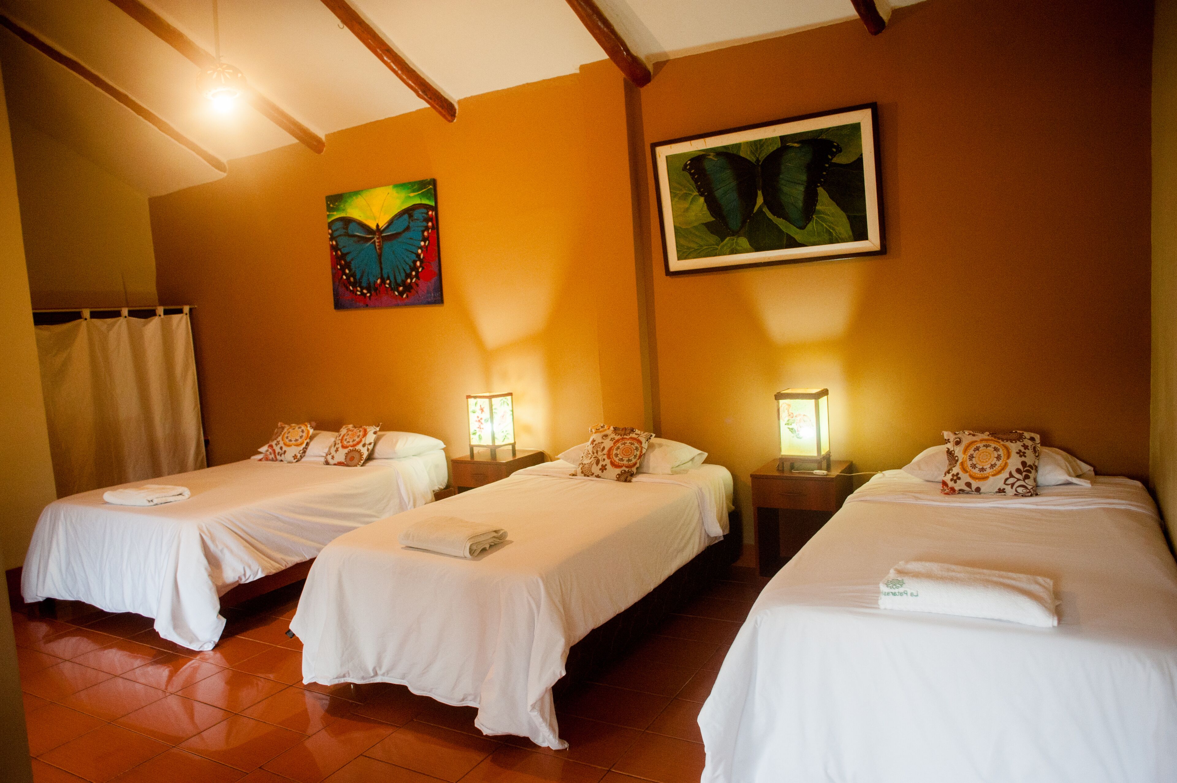 Habitación de hotel con tres camas individuales, cada una con edredón y almohadas blancos a juego. Hay dos mesitas de noche con lámparas, y el suelo está cubierto de baldosas.