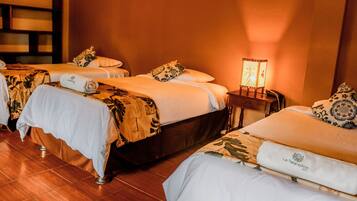 A hotel room with three beds, each with a white bedspread and patterned pillows. There is a small table with a lamp between the beds.