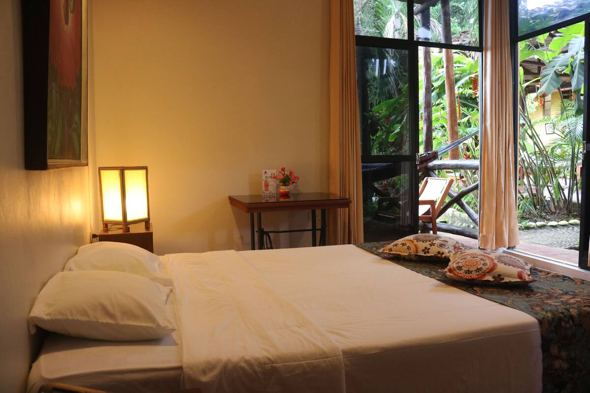 A hotel room with a bed, a small table with a lamp, and a view of greenery through a window.