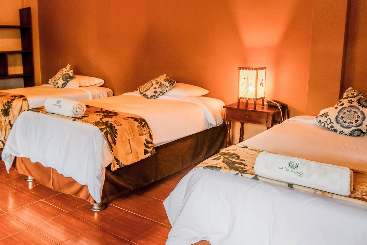 A hotel room with three beds, each with a white bedspread and a floral-patterned blanket. There is a nightstand with a lamp between the beds.