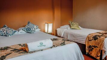 Habitación de hotel con dos camas, cada una con una almohada y una manta. Hay una mesita de noche con una lámpara entre las camas.