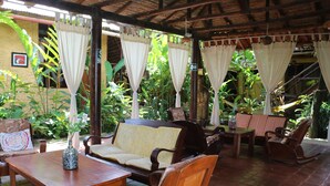 An outdoor patio with wooden furniture, including a sofa and chairs, surrounded by lush greenery and a thatched roof structure. - La Patarashca Hotel (Tarapoto)