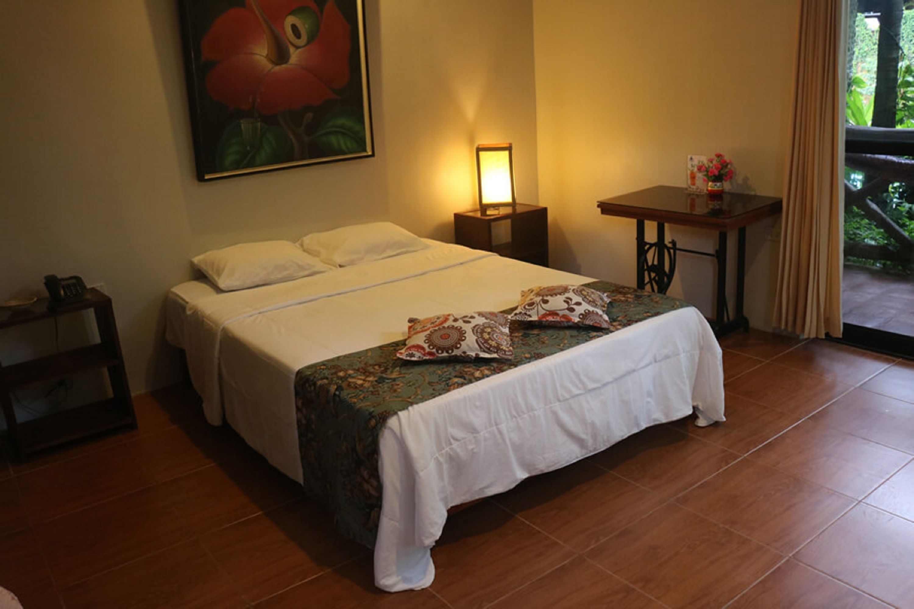 Un lit bien fait, recouvert d’une courtepointe blanche et agrémenté de coussins décoratifs, une table de chevet avec une lampe, et un tableau représentant une fleur accroché au mur.