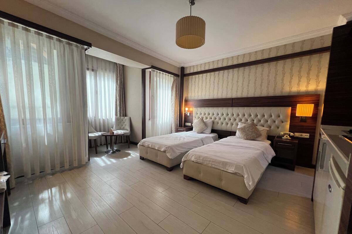 A hotel room with two beds, a sitting area with a chair and table, and a large window with curtains.
