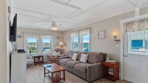 Ein gemütliches Wohnzimmer mit Sofa, Sessel und Couchtisch. Es gibt Fenster mit Vorhängen, einen Deckenventilator und eine Tür mit Blick ins Freie.