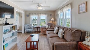 Ein Wohnzimmer mit Deckenventilator, Fernseher, Bücherregal, braunem Sofa, gemustertem Sessel und Blick ins Freie.