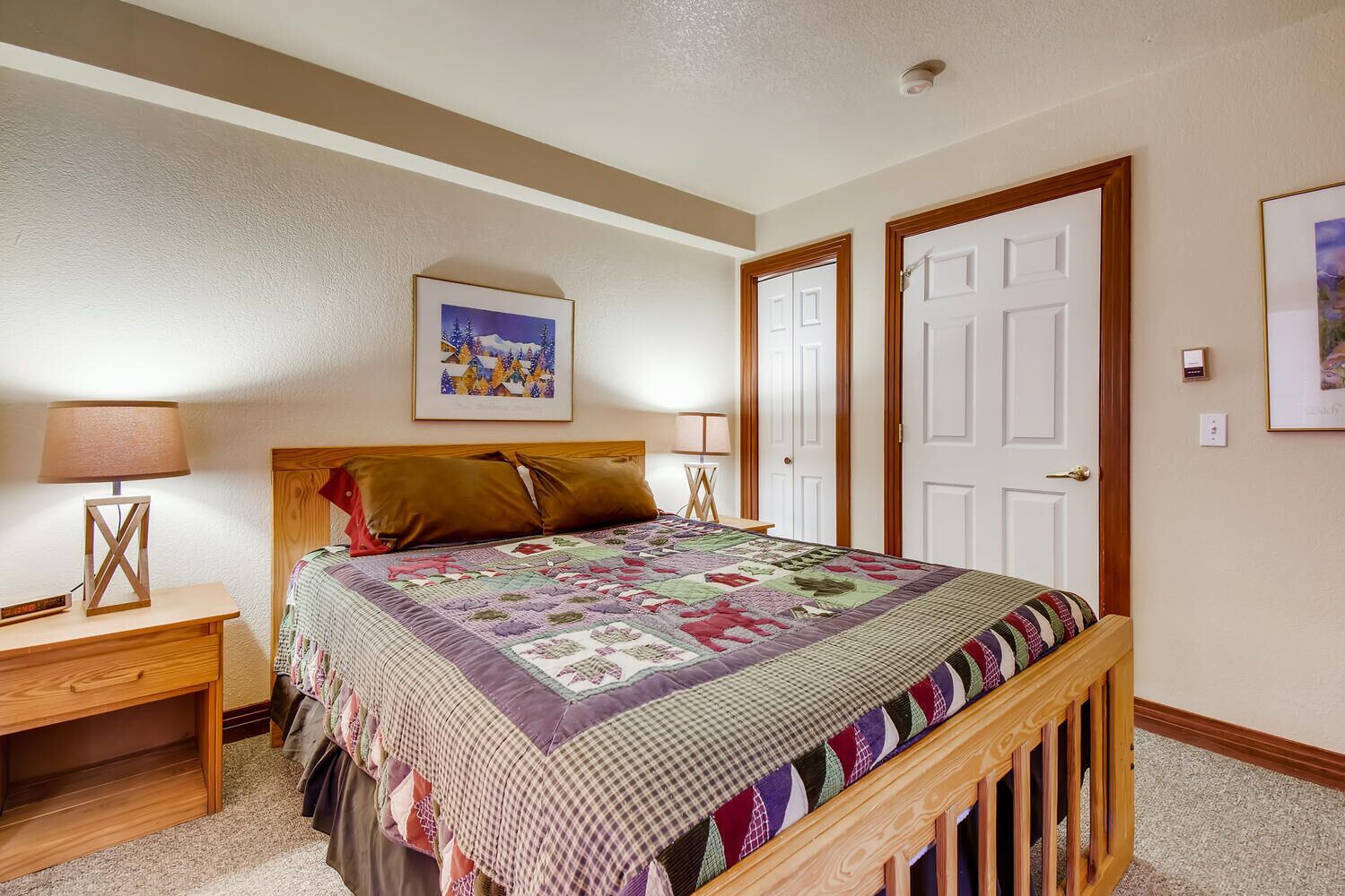 Un dormitorio con una cama de madera, una mesita de noche con lámpara, un cuadro en la pared y dos puertas.