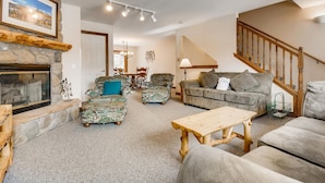 A cozy living room with a stone fireplace, a wooden coffee table, two patterned armchairs, a grey sofa, and a staircase. - Pinecreek #F - 3 BR: Close to Town- Shuttle to Slopes- Pool & Hot Tub Access (Breckenridge)