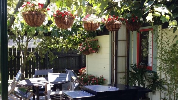 An outdoor patio with tables and chairs, hanging flower baskets, and a window with red trim.