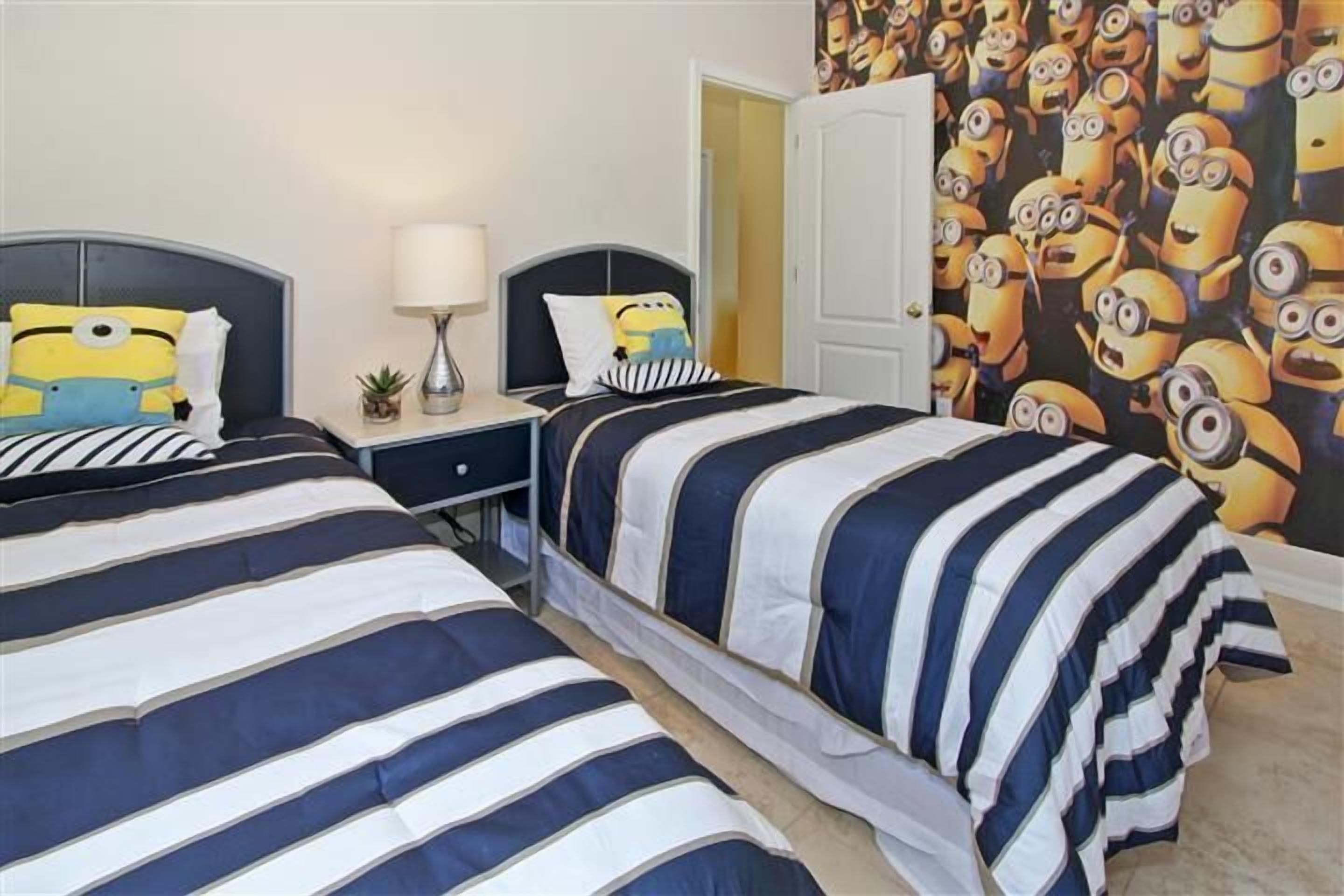 A hotel room with two beds, striped bedding, a nightstand with a lamp, and a Minion-themed wall.