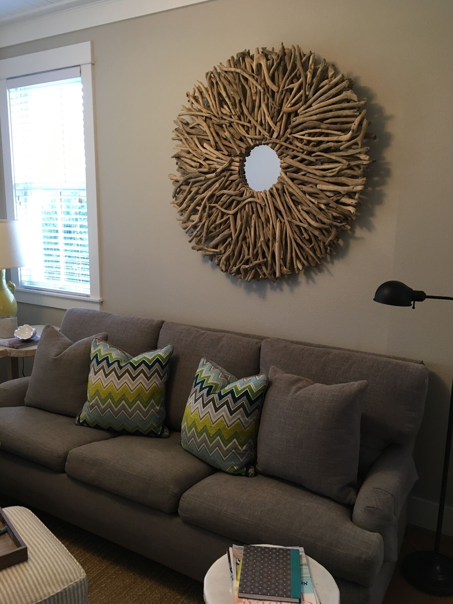 Un salon avec un canapé gris, deux coussins à motifs, un élément décoratif rond fixé au mur, une petite table d’appoint avec une lampe et un lampadaire.