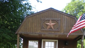 A wooden cabin with a porch, two chairs, a door, and an American flag. - Tiny house with full-size amenities tucked away in the countryside (Sulphur)