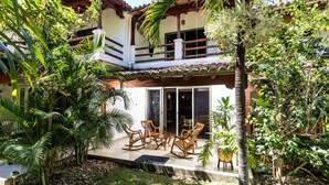 A two-story house with a balcony, a patio with wooden chairs, and lush greenery. - Town House 9: Comfort and golf course view (Hacienda Iguana)