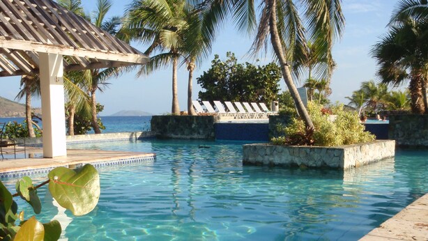 A clear pool with a tiled deck, surrounded by palm trees and a view of the ocean. - Corner Villa with Stunning Sapphire Bay Views – Sleeps 6 w/Backup Generator. (St Thomas)