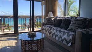 A room with a large window, a glass table, a sofa, and a view of the ocean. - Corner Villa with Stunning Sapphire Bay Views – Sleeps 6 w/Backup Generator. (St Thomas)