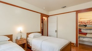 Un dormitorio con dos camas, cabeceros de madera, una mesita de noche con lámpara, un armario con ropa doblada y una puerta que da a otra habitación.