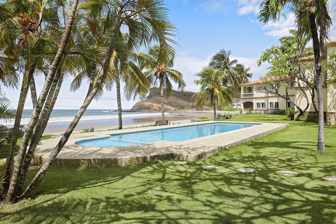 A swimming pool surrounded by palm trees, a beach, and a building with a balcony overlooking the ocean.