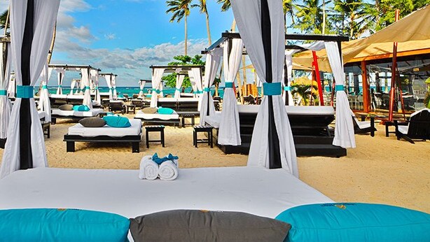 Um resort à beira-mar com várias espreguiçadeiras com toldos, algumas com almofadas e cobertores, dispostas na areia.