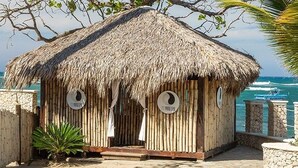 Uma cabana com telhado de palha, parede de pedra e cerca de madeira, situada perto de uma praia com o oceano ao fundo.