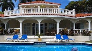 Uma casa de dois andares com piscina, espreguiçadeiras azuis e um espaço de varanda coberto.
