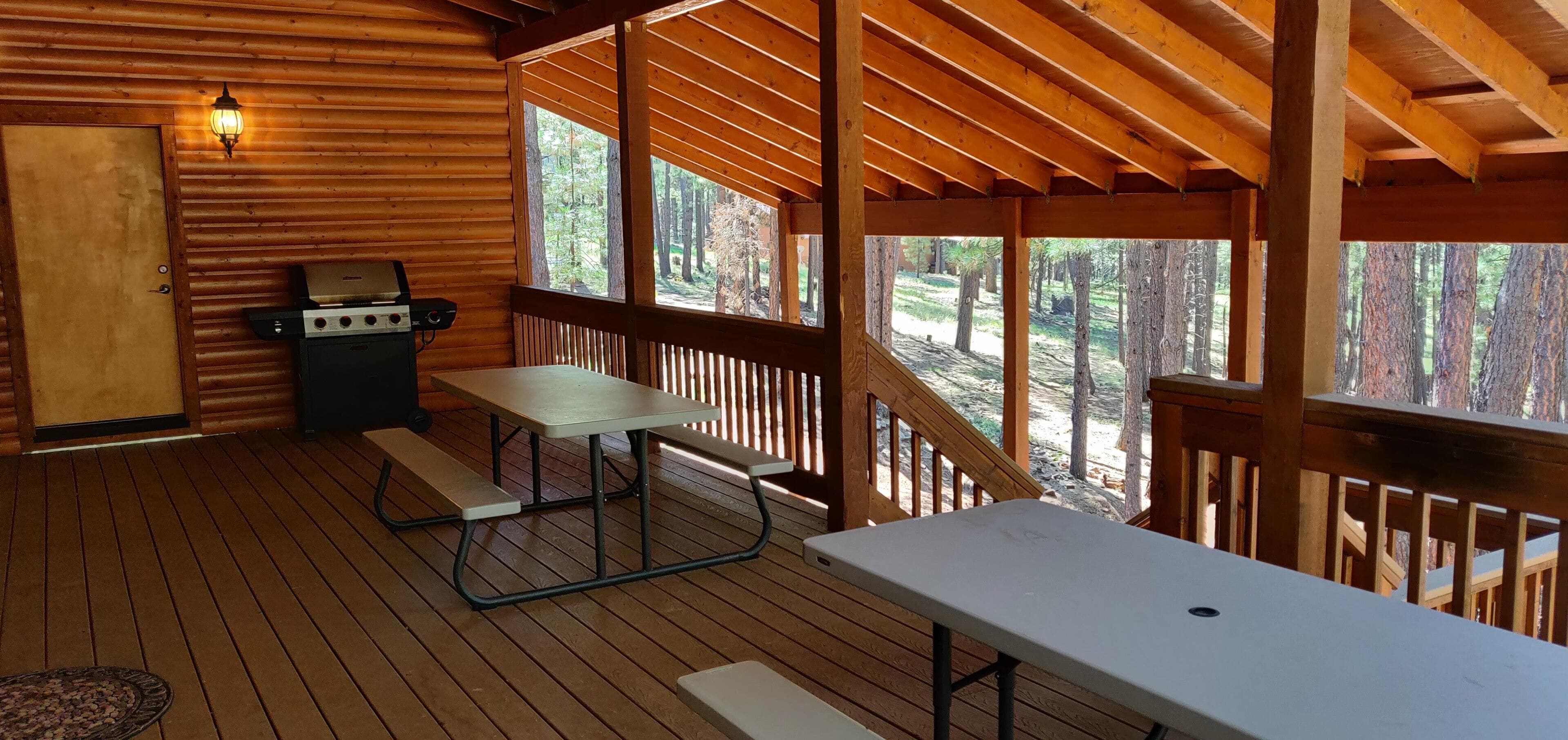 A wooden deck with picnic tables and a grill, overlooking a forested area.