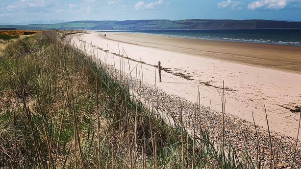 Ein Strand mit einem Zaun und Gras im Vordergrund, ein sandiger Strand und der Blick auf das Meer und ferne Hügel.
