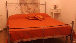 Un lit simple avec un cadre en métal et un protège-matelas rouge, une petite table de chevet avec une lampe, et un rideau à la fenêtre.
