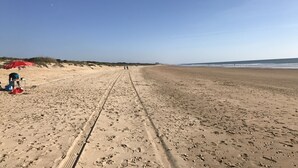 Une vaste plage de sable, parsemée d’empreintes de pas et de traces de pneus, un parasol rouge et quelques personnes visibles au loin.