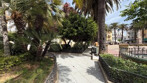 Un chemin pavé bordé de palmiers et agrémenté d’une balustrade en pierre, qui mène à une place où se trouvent une statue et des bancs.
