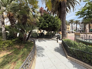 Un sendero pavimentado flanqueado por palmeras y una barandilla de piedra, que conduce a una plaza con una estatua y bancos.