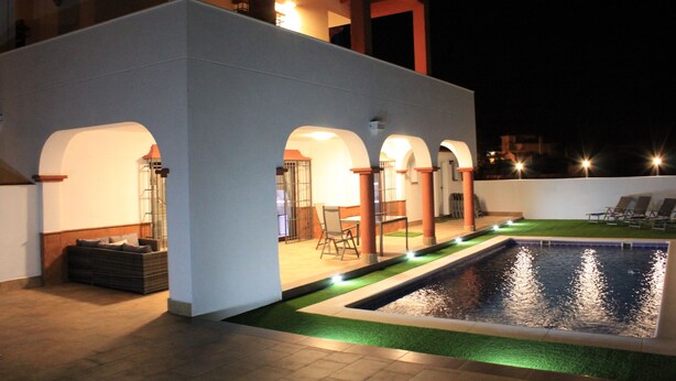 Un espace piscine comprenant une piscine rectangulaire, un coin salon couvert avec des chaises, et un petit jardin avec une pelouse et un élément décoratif aquatique.