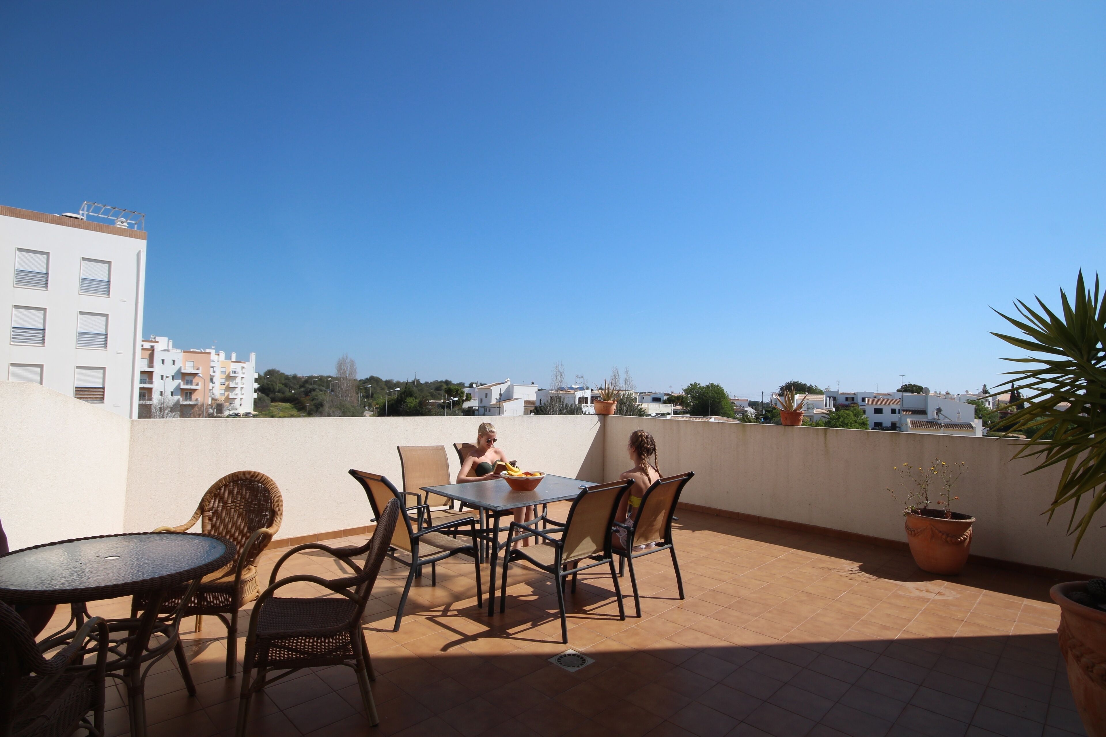 A rooftop terrace with a table and chairs, potted plants, and a view of buildings in the distance.