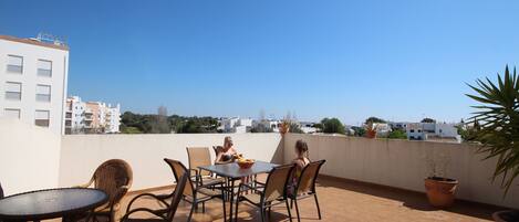 A rooftop terrace with a table and chairs, potted plants, and a view of buildings in the distance.