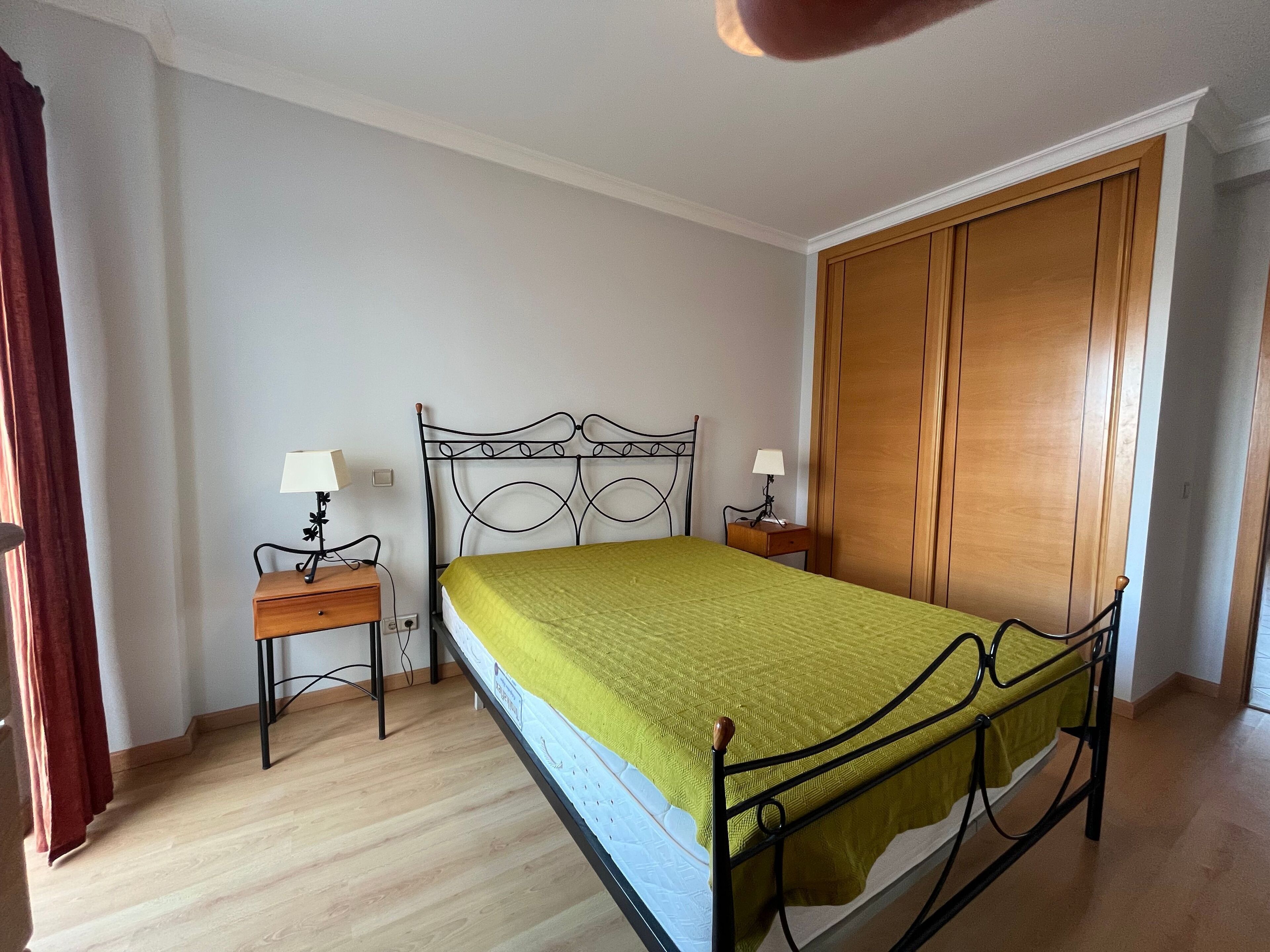 A bedroom with a bed, two bedside tables with lamps, a wooden wardrobe, and a wooden floor.
