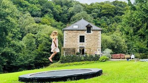 Una casa de pedra amb un pati en verd, un trampolí i una persona saltant.